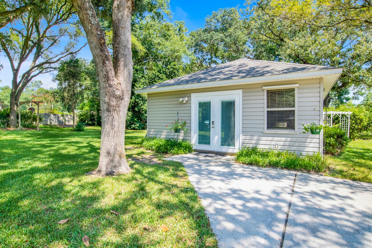 A charming guest house is positioned on a spacious lawn, surrounded by lush greenery and mature trees. A concrete pathway leads to double glass doors framed by plants, offering a welcoming entrance. The serene outdoor space is highlighted by well-maintained grass and a clear blue sky.