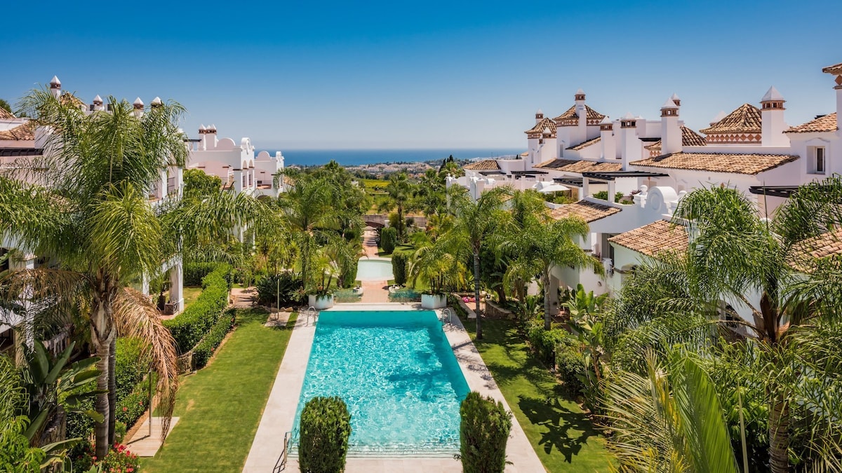 The image captures a serene communal area featuring a large swimming pool surrounded by lush greenery and palm trees. Whitewashed buildings with terracotta roofs are visible in the background, with the coastline and ocean extending beyond. Clear blue skies create a bright atmosphere.