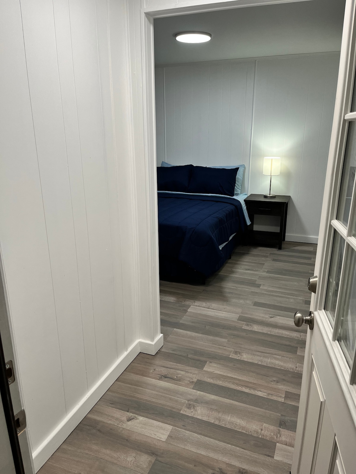 An inviting entryway leads into a cozy sleeping area, featuring a queen bed dressed in dark blue bedding. A bedside table with a lamp provides soft illumination, while light-colored walls and stylish flooring enhance the room's spacious feel.