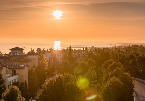 A stunning sunset casts warm orange hues across the Pacific Ocean, reflecting on the water. Lush greenery frames the scene, with buildings in the foreground blending into the natural landscape. The coastline and distant horizon are softly outlined in the fading light.