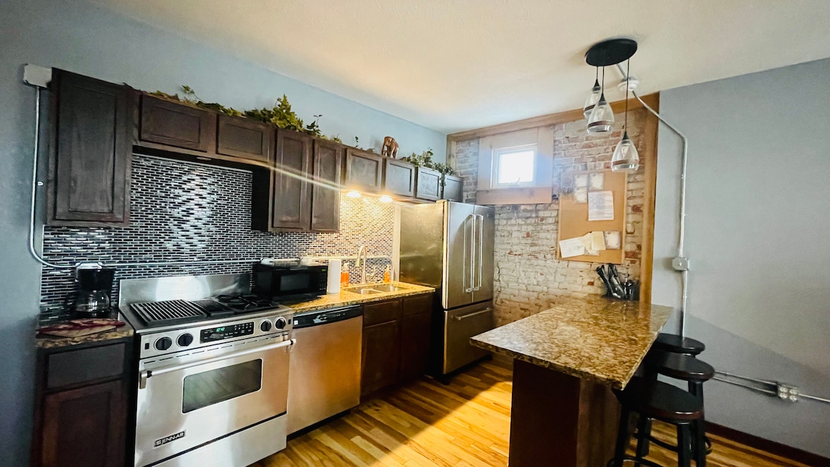 The kitchen features modern stainless steel appliances, including a gas stove and refrigerator. A granite countertop runs along one side with three dark wooden stools positioned nearby. Exposed brick walls add character, while warm lighting creates a welcoming space.