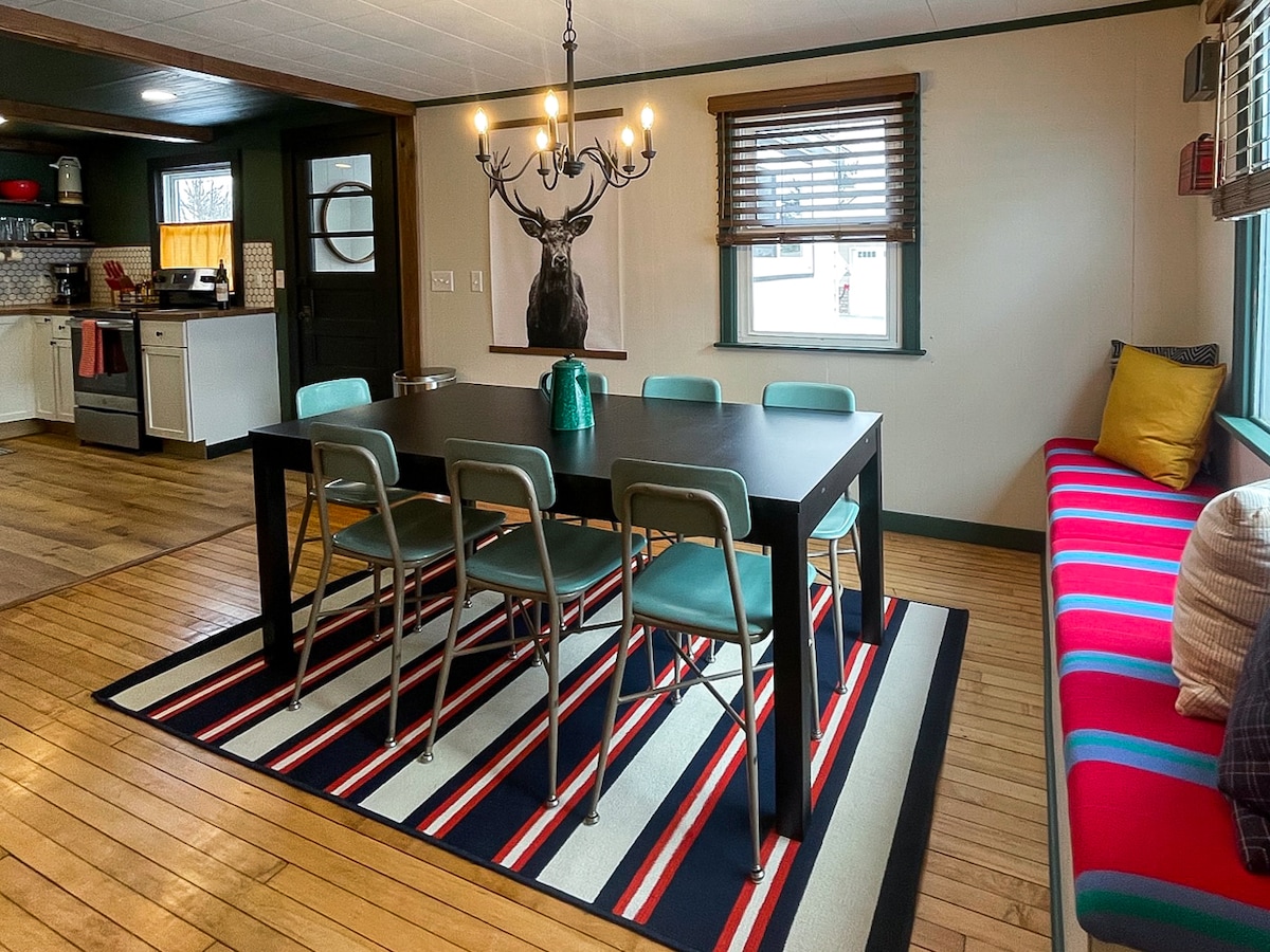 A spacious dining area features a large black table surrounded by six teal chairs, placed on a striped rug. A vibrant window seat adds color, while a chandelier hangs above, contributing to the room's inviting ambiance. The kitchen can be seen in the background.