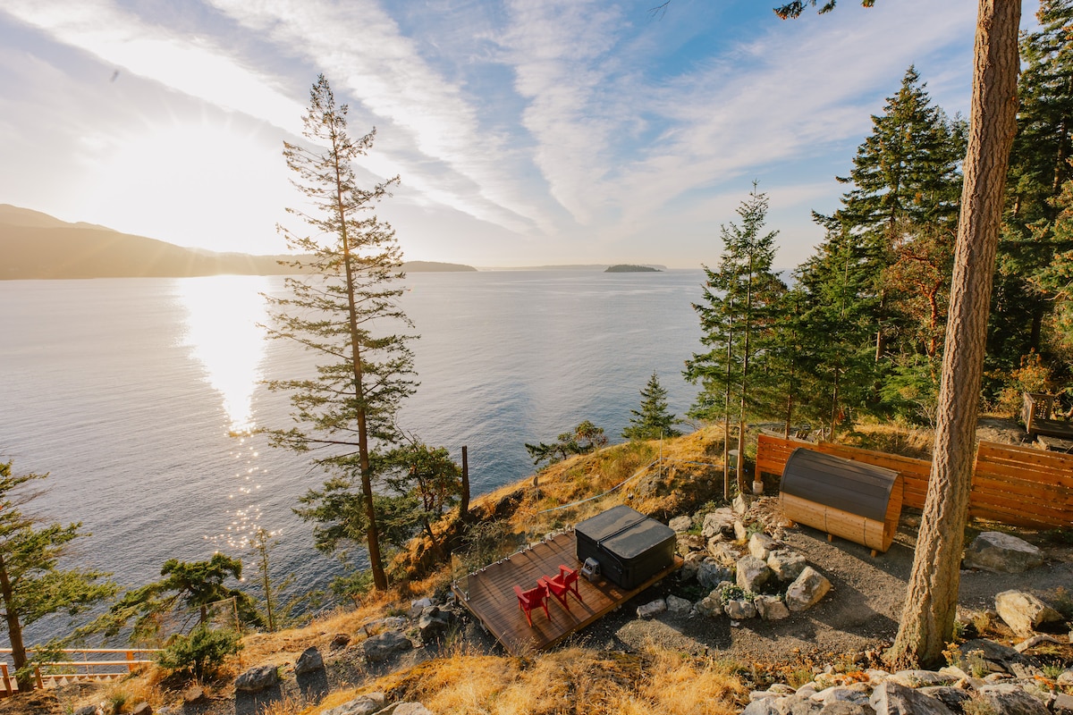 A serene outdoor space features a hot tub and a wood barrel sauna, surrounded by natural stone and tall trees. The ocean glistens in the morning light, with hills visible in the distance under a clear sky. Cozy red lounge chairs are positioned nearby.