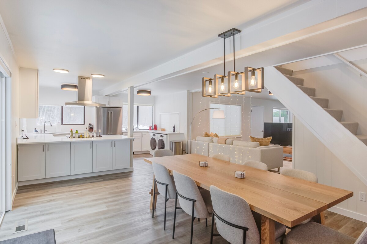 An open-plan dining and kitchen area is highlighted by a large wooden dining table surrounded by upholstered chairs. Modern lighting fixtures hang above, and an adjacent kitchen features sleek cabinetry and appliances. A staircase is visible in the background, leading to an upper level.