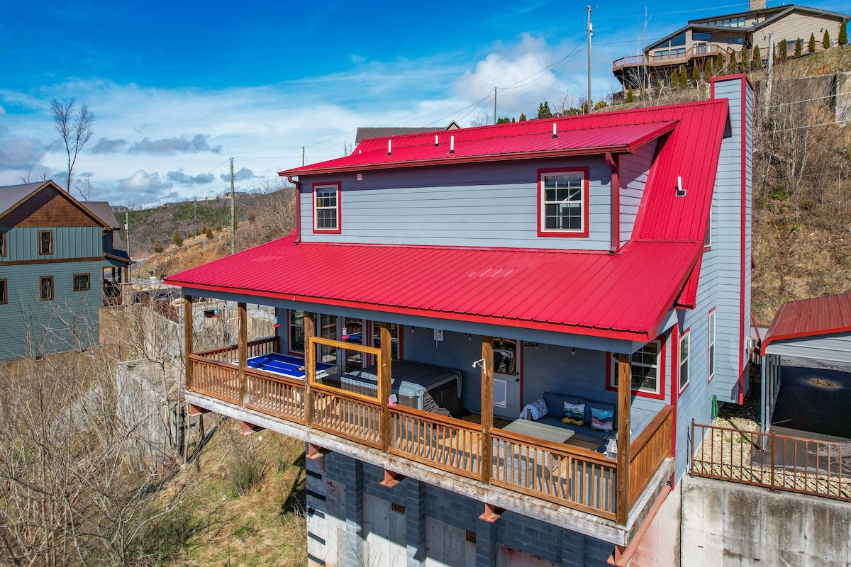 The exterior of the cabin showcases a vibrant red metal roof and light grey siding. A spacious front porch extends across the house, featuring wooden railings and outdoor seating. The surrounding area includes natural landscape views and nearby houses, enhancing the cabin's rustic charm.