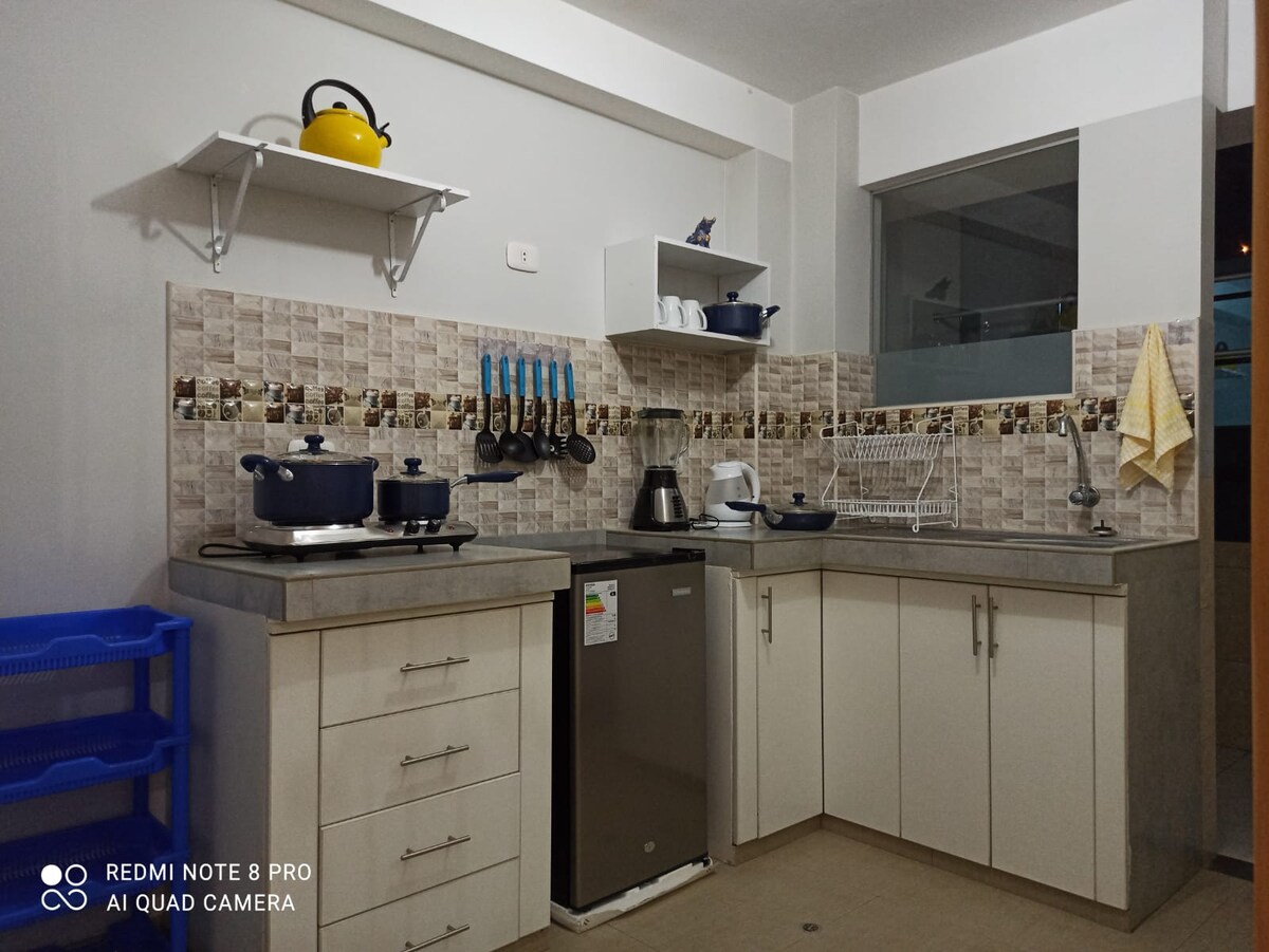 The kitchen features a modern design with light-colored cabinetry and contrasting stone countertops. Essential appliances like a stove, kettle, and refrigerator are visible. Kitchen utensils are neatly organized on the wall, and a window allows natural light to enter the space.