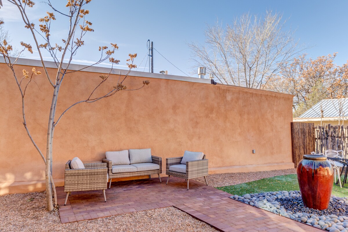 An outdoor space features a simple arrangement of woven seating beside a decorative pot on an area of pebbles. A tree provides subtle shade, complementing the adobe wall in the background. A patch of grass is visible, enhancing the cozy atmosphere.