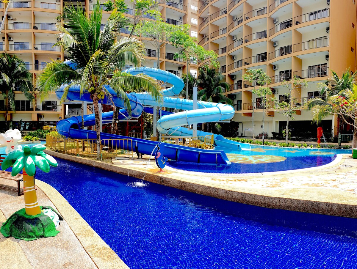 The water theme park features a vibrant blue water slide curving above a pool area, surrounded by lush greenery and tropical plants. A playful palm tree sculpture stands in the foreground, while the multi-story resort buildings are visible in the background.