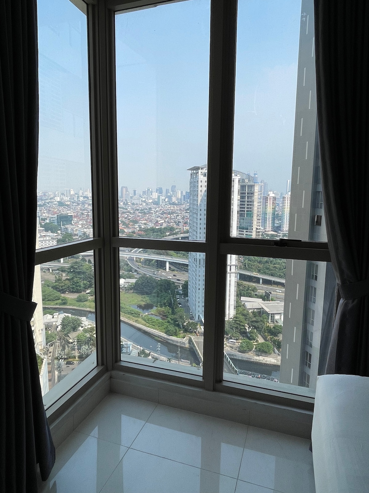 A bright corner window offers a panoramic city view, showcasing urban skyscrapers and roads below. White curtains frame the glass, allowing natural light to illuminate the interior space.