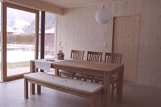 A dining area is shown with a large wooden table and five matching chairs. A bench with a padded seat is positioned alongside the table. Natural light enters through a large glass door, revealing a view of the outdoor space.