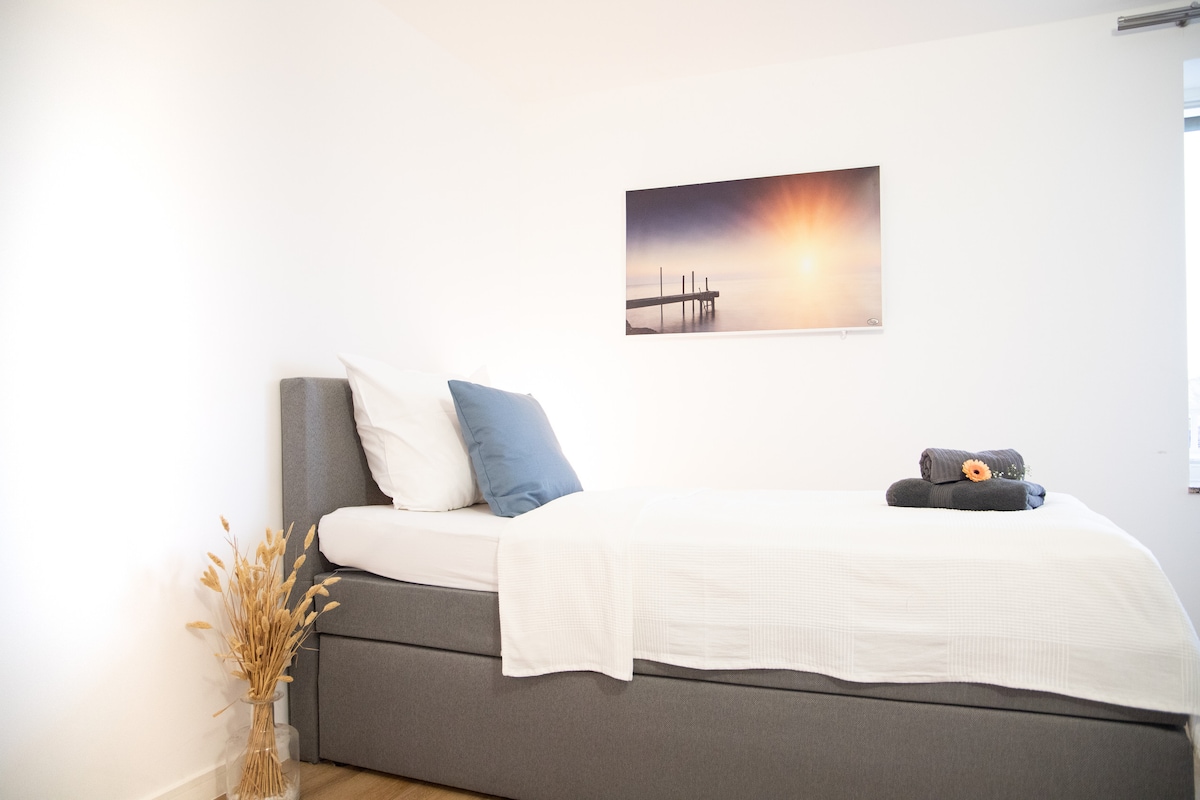 A spacious bed is positioned against a bright wall, topped with white linens and decorative pillows. A simple bouquet of dried grass adds a touch of natural decor beside the bed. A landscape artwork depicting a serene sunrise is hung above, enhancing the calming ambiance.