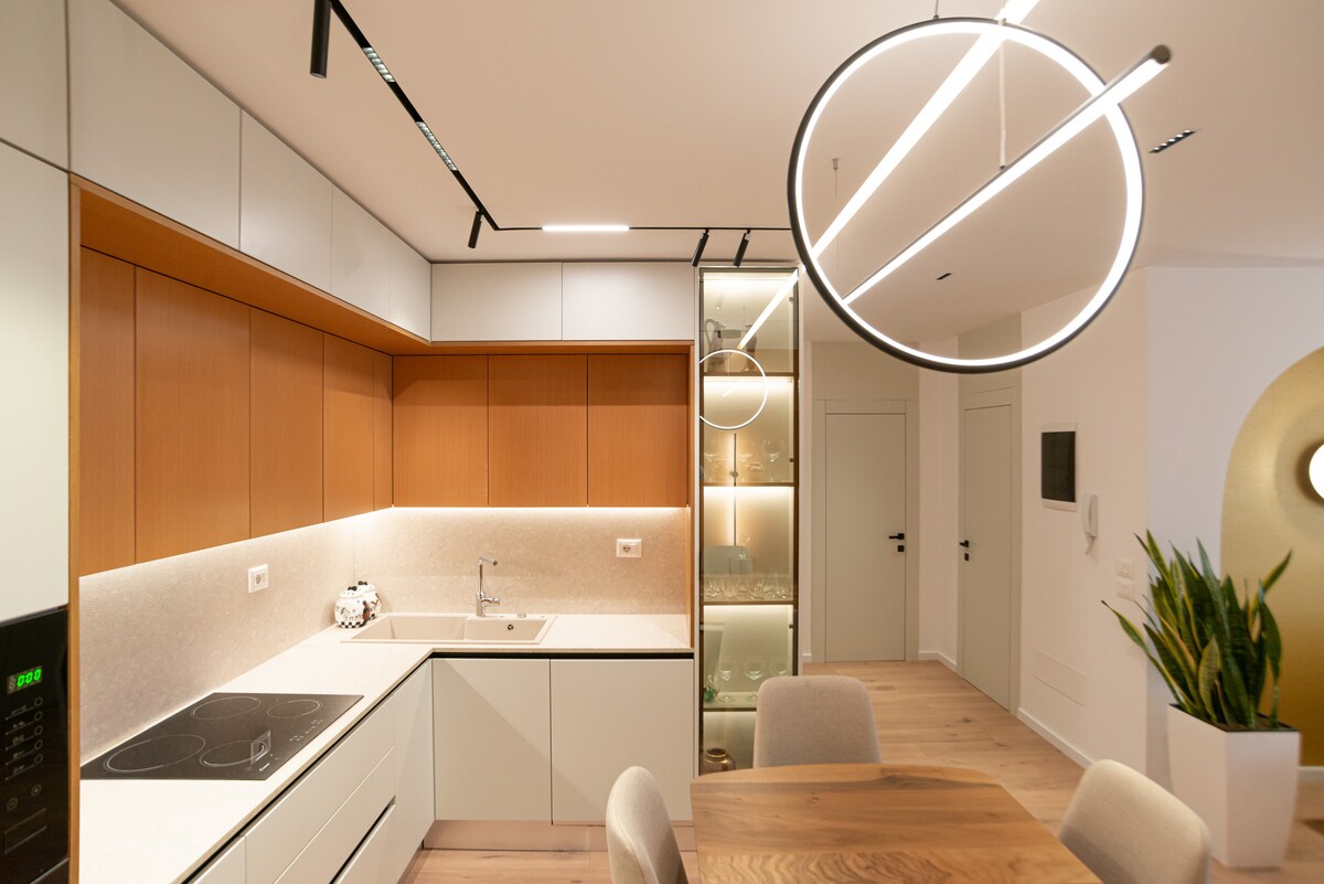 A modern kitchen features sleek cabinetry in warm wood tones, highlighted by under-cabinet lighting. A minimalist island with a stovetop is complemented by a dining table and softly glowing circular pendant lights. A contemporary display cabinet showcases decorative items.