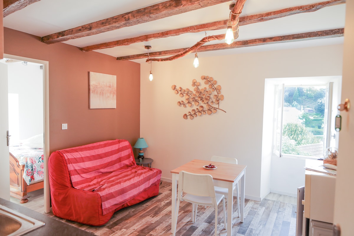 A living area features a bright red sofa with a striped pattern and a small wooden dining table surrounded by white chairs. Natural light enters through a window, illuminating the space. Decorative elements include a wall with wooden beams and an artwork mounted above the sofa.