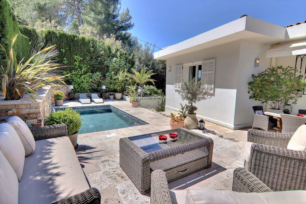 An outdoor seating area features comfortable sofas and a coffee table, surrounded by lush greenery. A private pool is visible nearby, with lounge chairs positioned elegantly beside it. The space is designed for relaxation and enjoyment in a serene setting.
