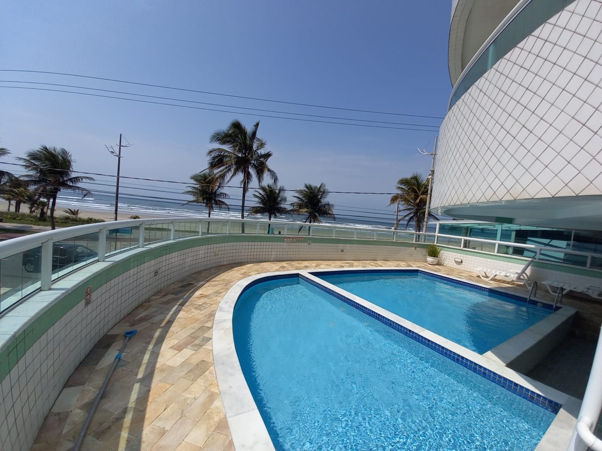 A swimming pool is seen set against an ocean view. Palm trees line the background, providing a tropical feel. The pool area is surrounded by light-colored tiles, and the building's modern architecture features a smooth, curved design.