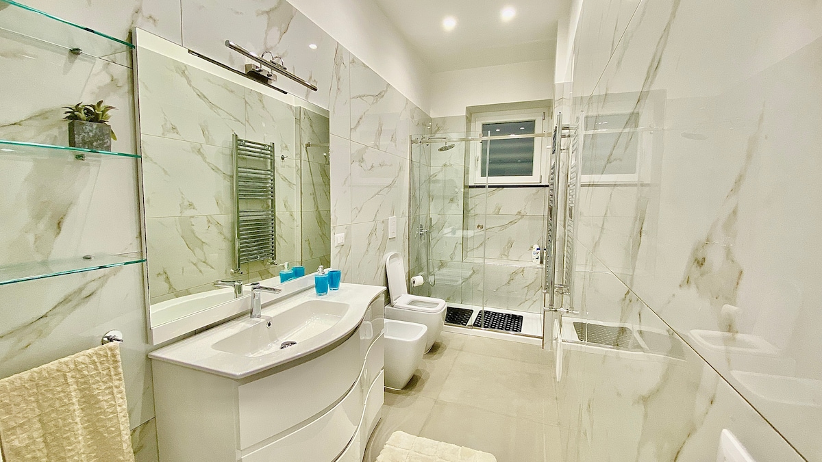 A modern bathroom features elegant marble walls and a sleek double vanity with a curved design. A large mirror above the sink reflects natural light, while a frosted window provides privacy. Ample storage is available on glass shelves, and a soft rug adds comfort underfoot.