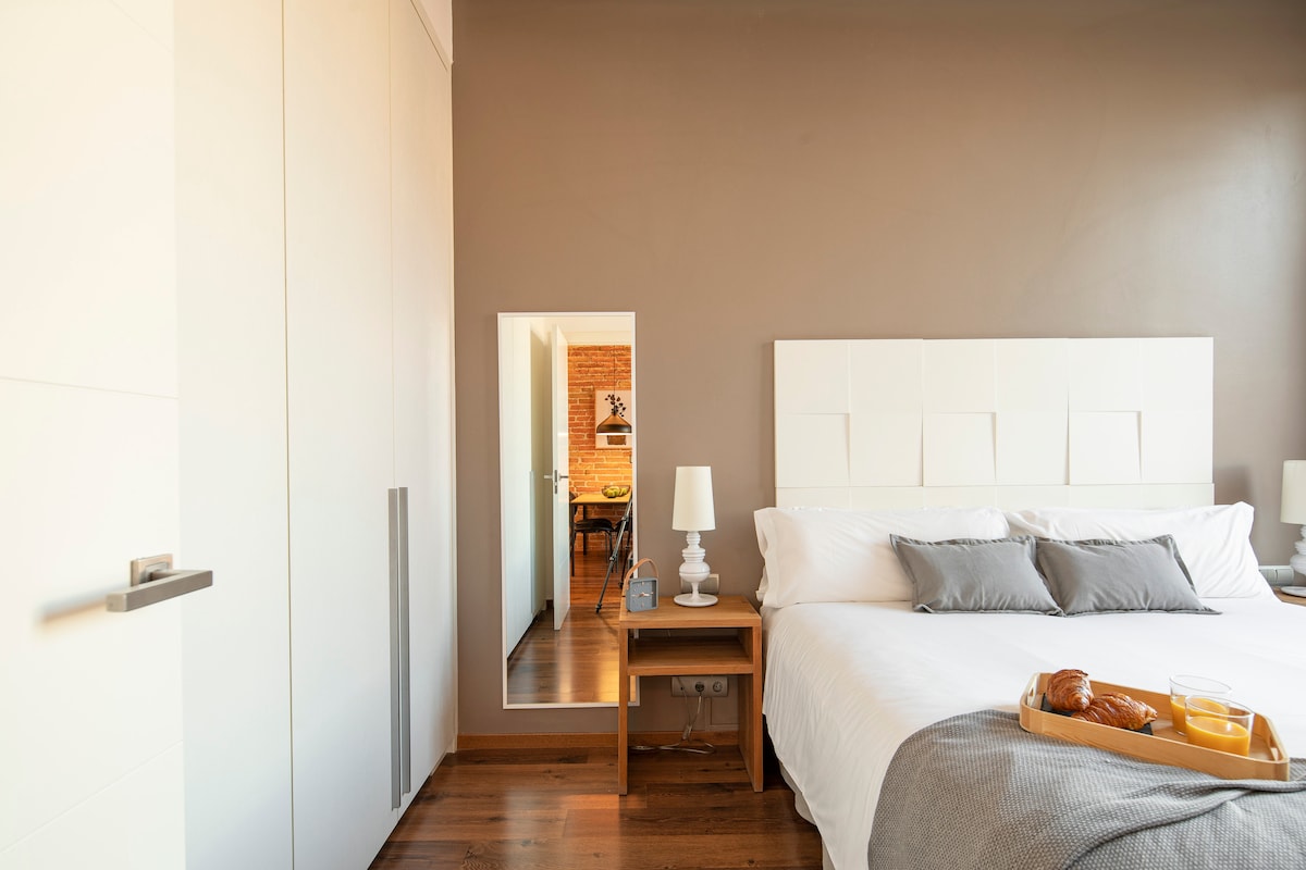 A cozy bedroom features a spacious bed with neutral linens and decorative pillows. A bedside table with a lamp is situated next to the bed. A large closet is present, and a mirror reflects the adjoining dining area visible through an open door.