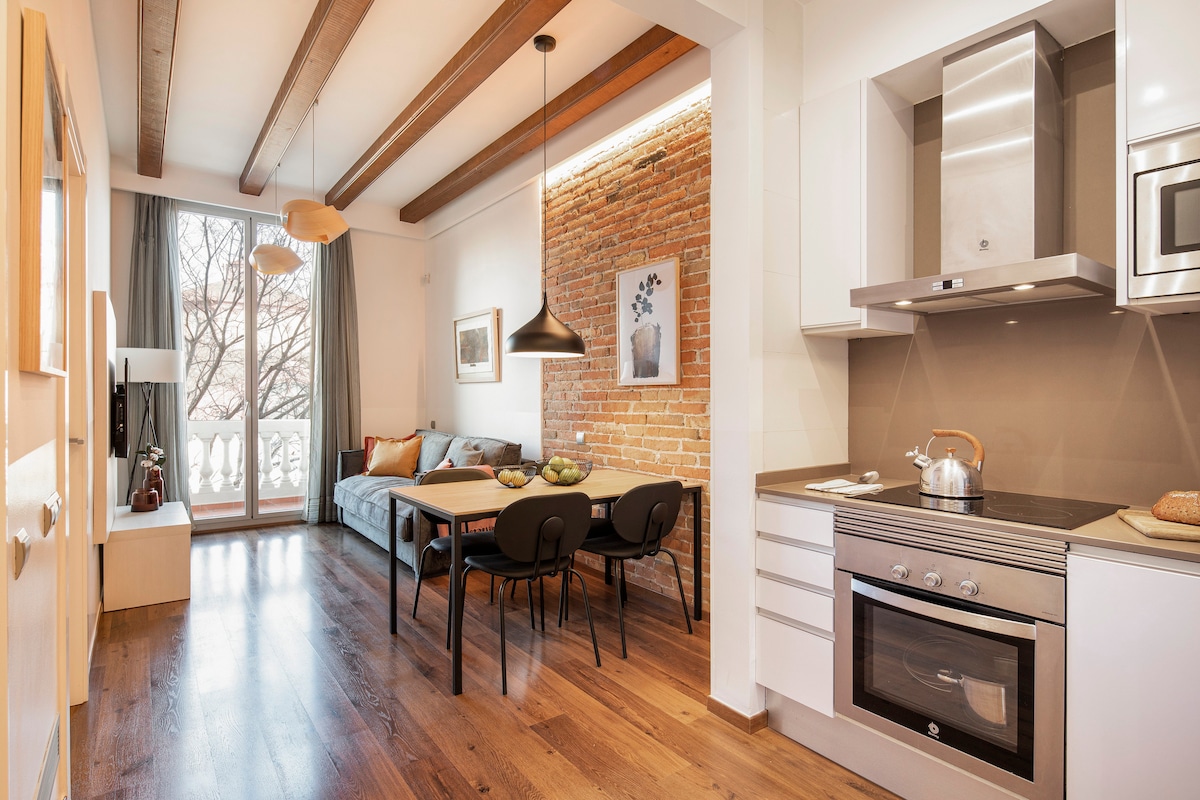 An inviting living area features a dining table with black chairs, complemented by a relaxing sofa. A rustic brick wall adds character, while large windows allow natural light to fill the space. Modern appliances, including an oven and kettle, are visible in the kitchen area.