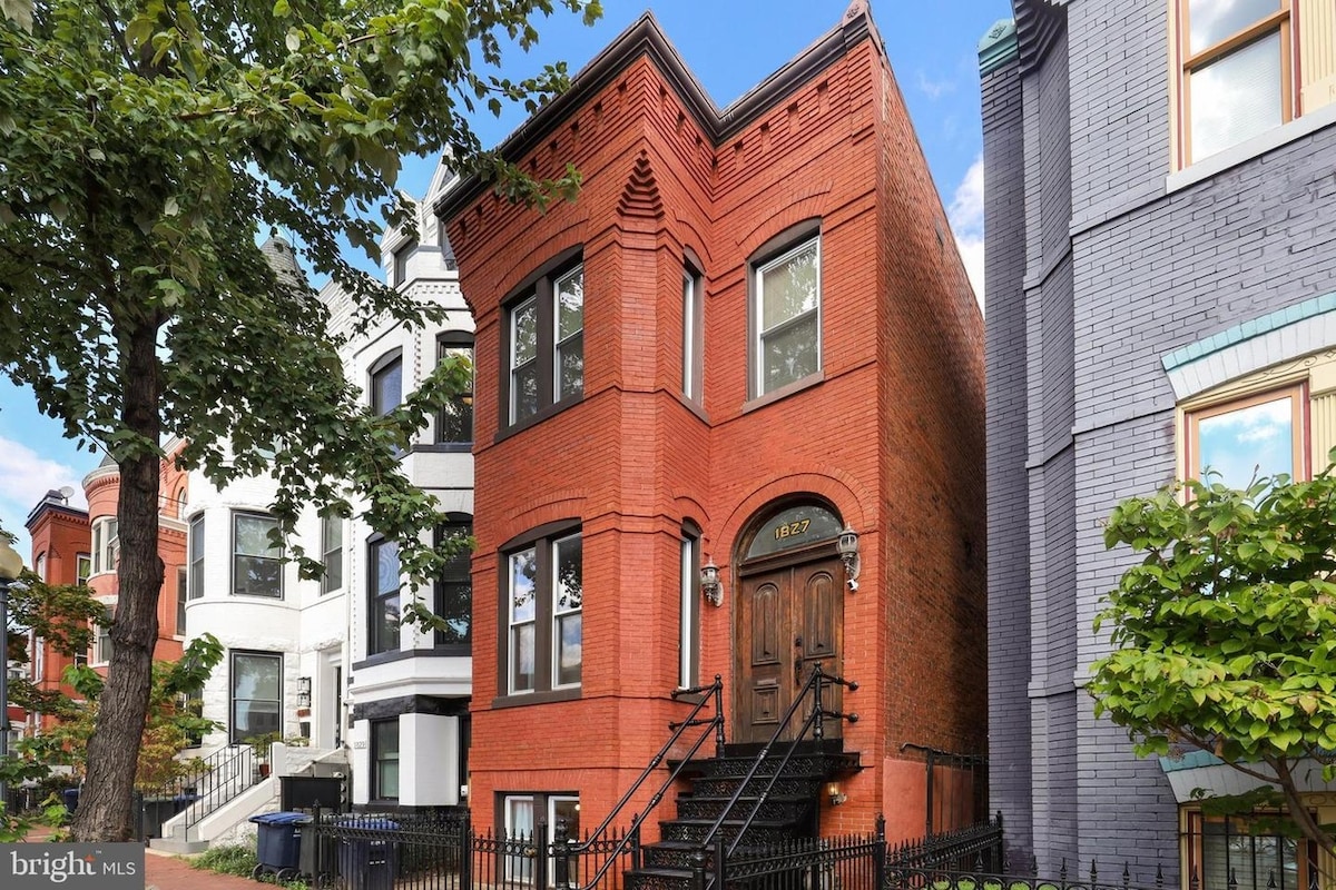 The exterior of a two-story townhouse is showcased, featuring a striking red brick facade with prominent architectural details. The entrance is framed by a large wooden door, flanked by symmetrical windows. Lush greenery from nearby trees complements the urban setting.