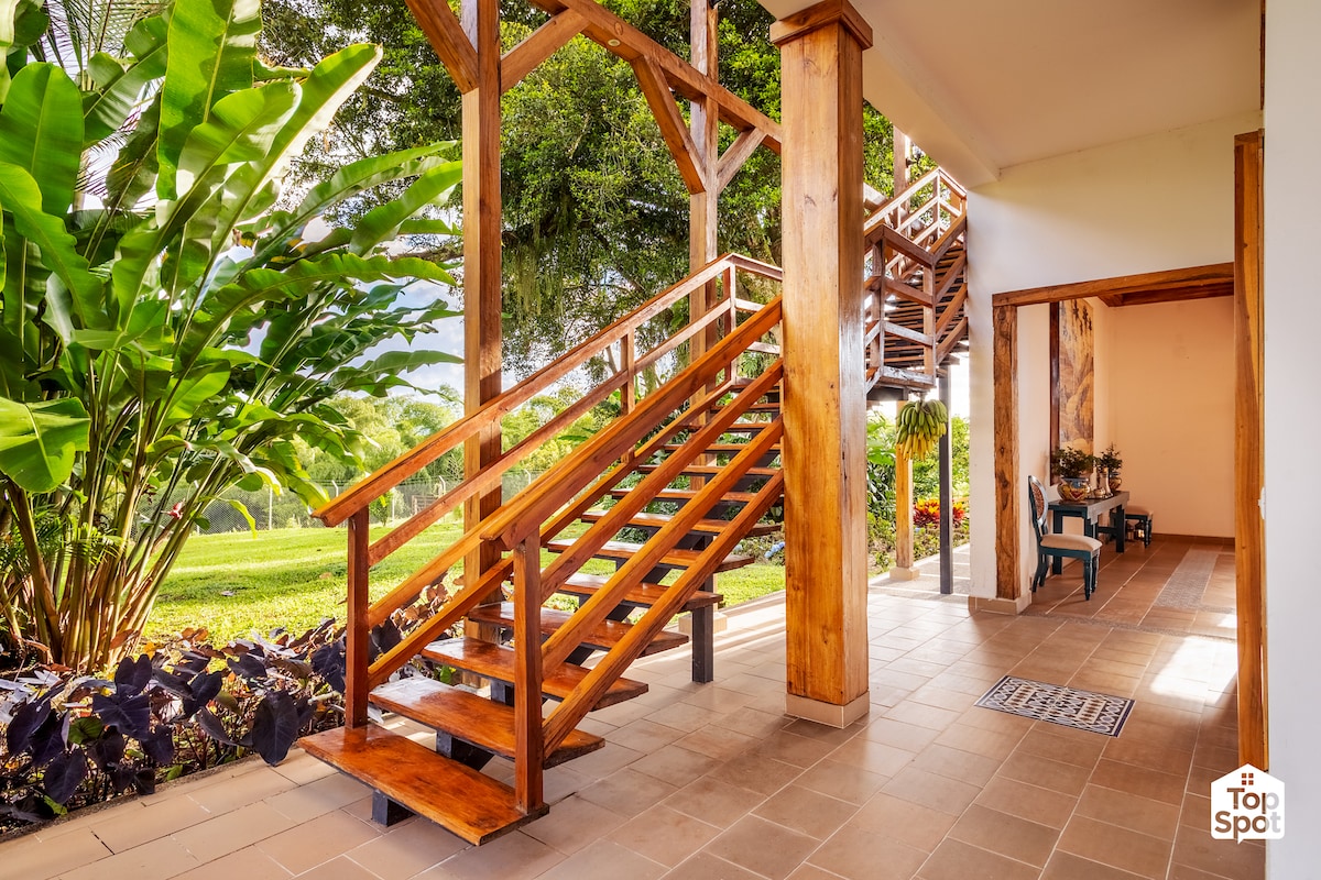 A well-maintained entrance features a wooden staircase leading to the upper level, surrounded by lush tropical plants. The flooring is tiled, and natural light illuminates the space, creating an inviting atmosphere. Seating areas are positioned nearby, providing a serene view of the garden.