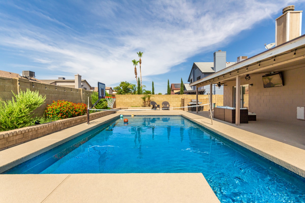 A spacious swimming pool is framed by a clean patio, with bright blue water reflecting the sky. Poolside seating is arranged for relaxation, and vibrant landscaping is visible along the edges. A nearby covered area houses an outdoor television, enhancing the recreational space.