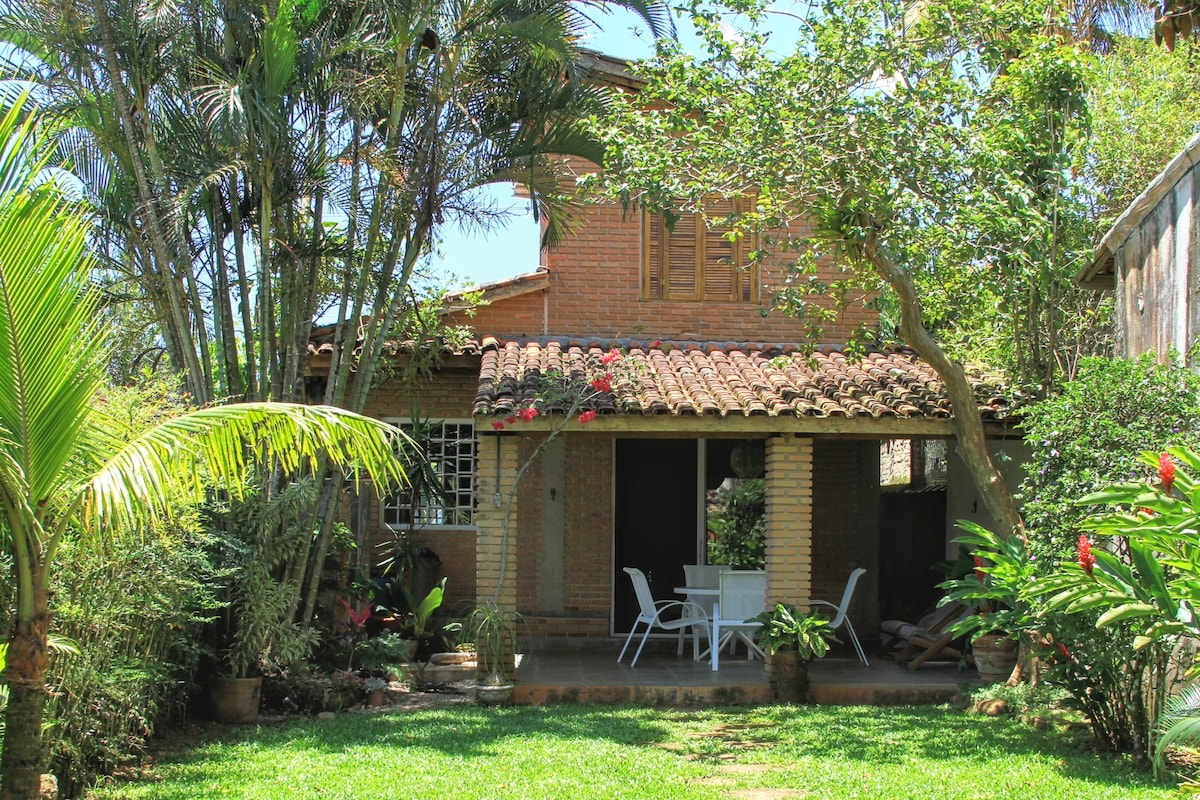 A two-story house is surrounded by lush greenery and palm trees. The exterior features a traditional tile roof and a welcoming entryway with a small patio area, including a white table and chairs. Vibrant flowers and plants add to the home's natural charm.