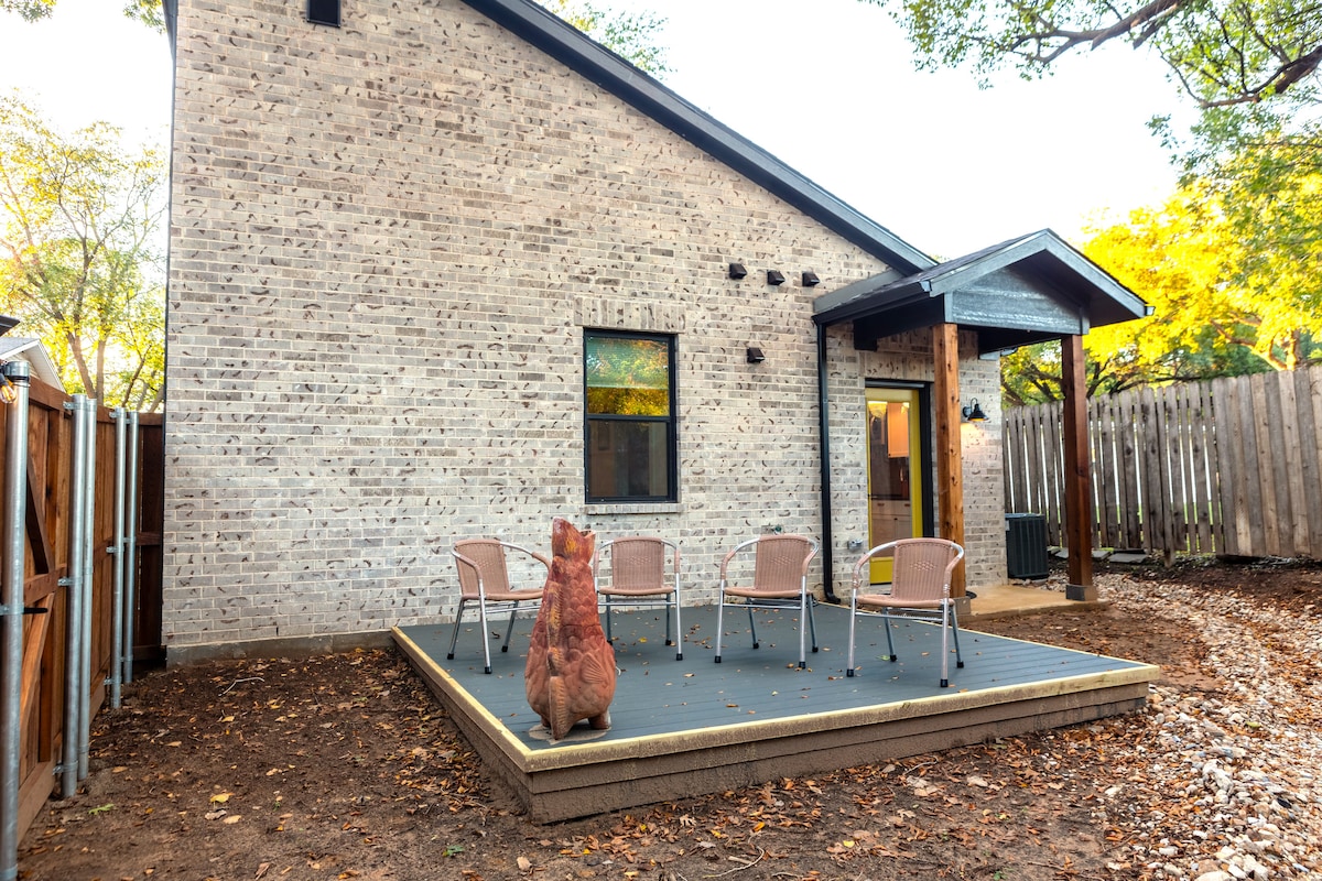 A private outdoor patio is featured, equipped with four metal chairs arranged around a central decorative element. The wooden deck is surrounded by trees, and the brick wall of the casita provides a modern backdrop. Natural light filters through the nearby window.