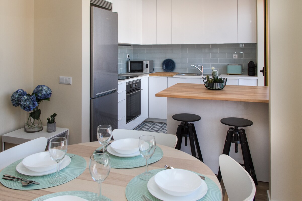 A modern kitchen is displayed, featuring sleek white cabinetry and a wood-topped island with two stools. The dining area showcases a round table set with plates and glasses, complemented by a vase of blue flowers on the counter.