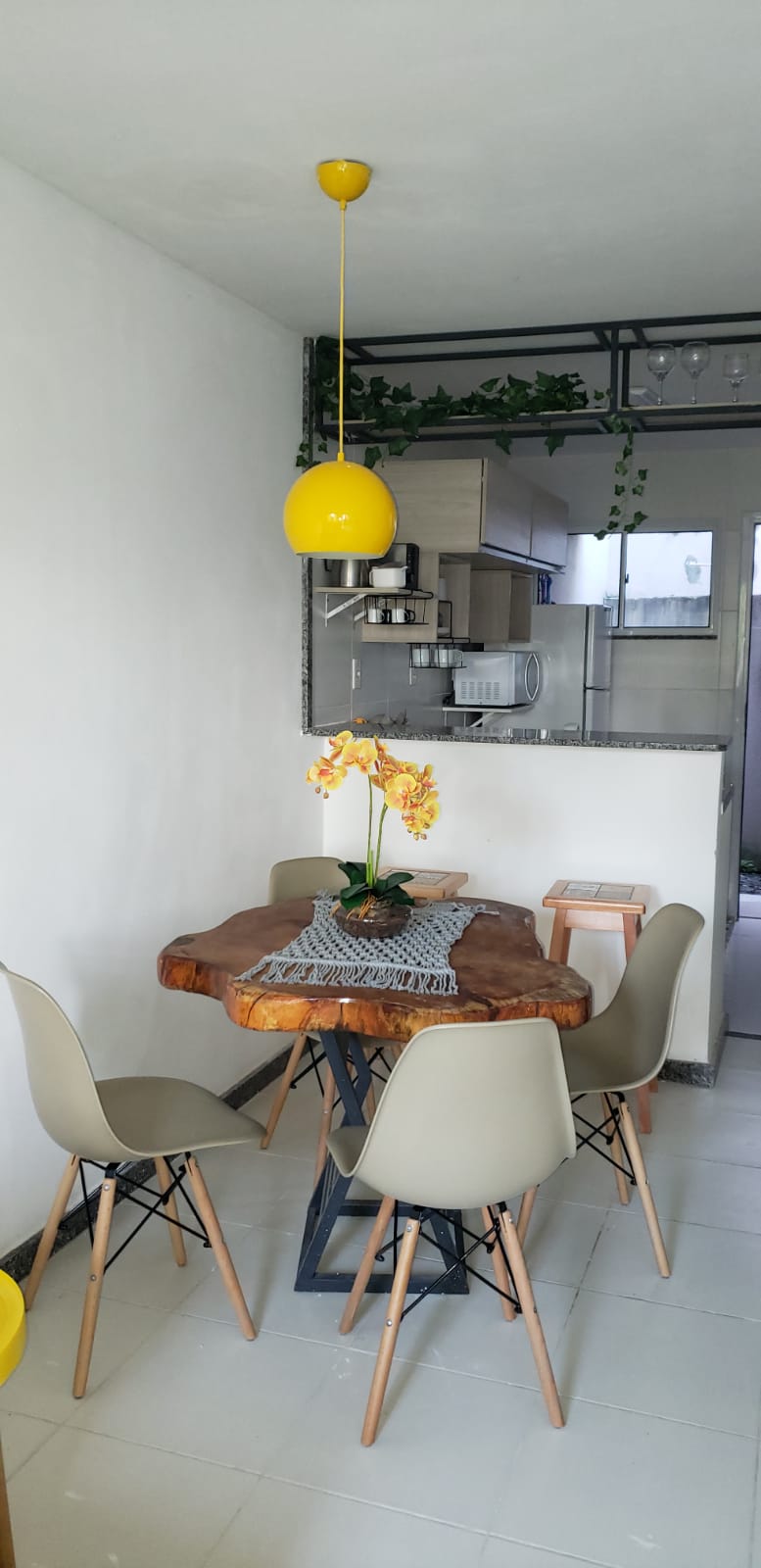 A dining area is presented with a wooden table featuring a natural edge, surrounded by four light grey chairs. A vibrant yellow pendant light hangs above, and a floral centerpiece adds a pop of color. The open kitchen area is visible in the background.