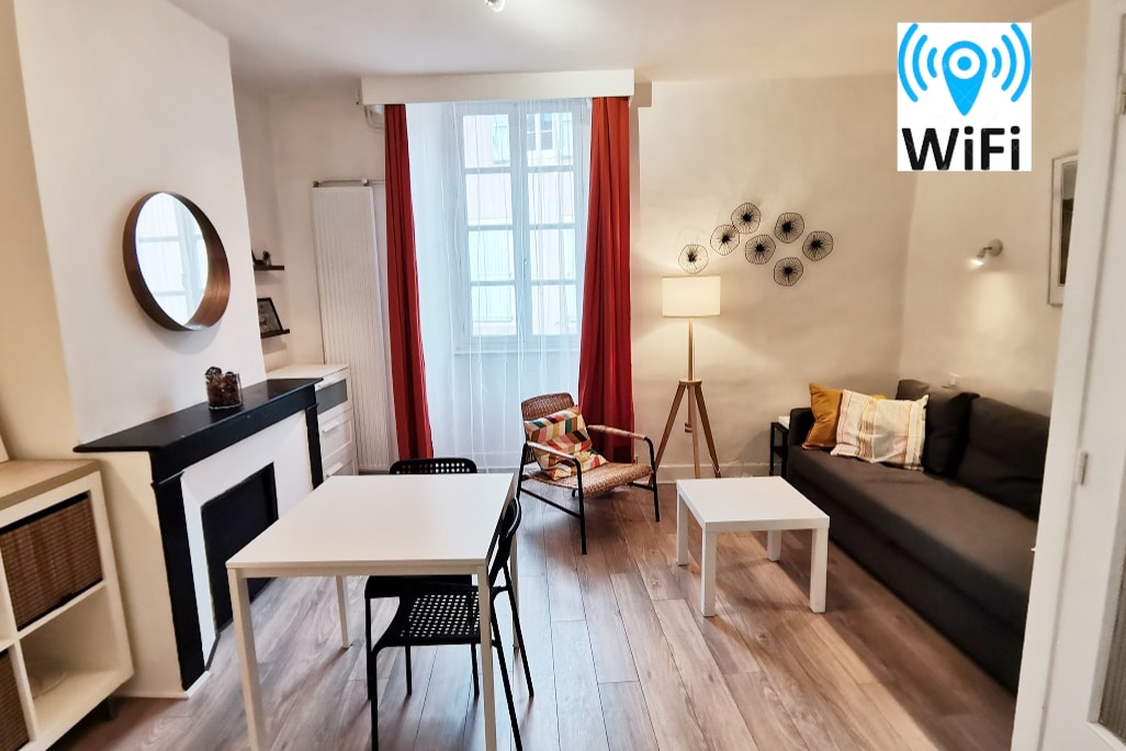 A modern, open living area is presented, featuring a gray sofa and a comfortable armchair beside a small dining table. A round mirror hangs above a decorative fireplace. Natural light filters through a window, complemented by soft curtains, enhancing the peaceful ambiance.