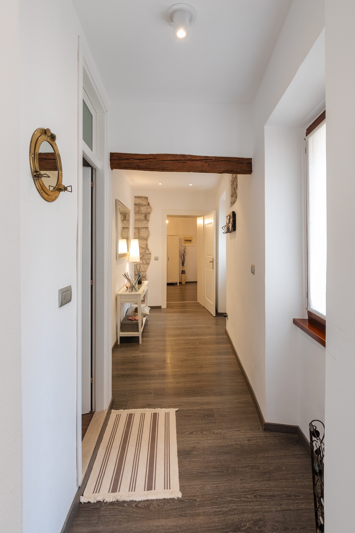 A narrow hallway is seen featuring smooth wooden flooring and white walls. A striped runner rug adds texture, while a small console table with decorative items is positioned against one side. Natural light enters through a window on the opposite end, enhancing the space.