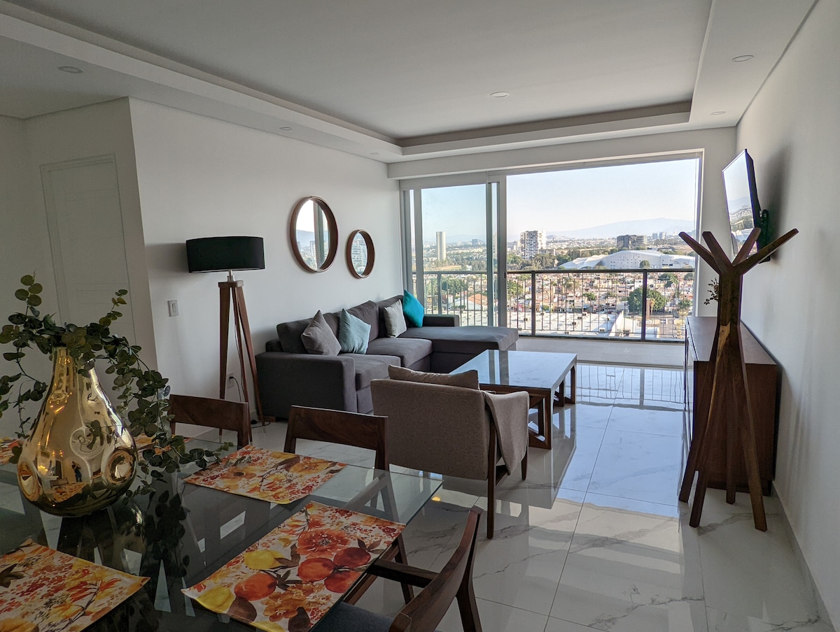 A bright living area features a comfortable sectional sofa and a flat-screen TV mounted on the wall. Large windows provide panoramic views of the surrounding cityscape, while a dining table is set with floral placemats. Decorative mirrors and plants add subtle touches.
