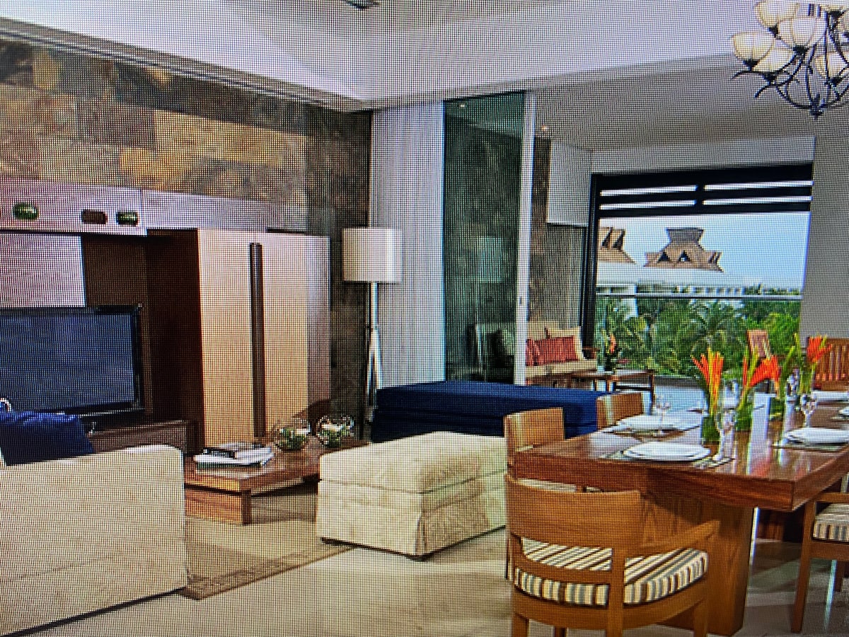 A modern living area features a neutral color palette with plush seating arrangements and a wooden dining table. Large windows provide natural light and views of the outdoors. Decorative elements include a television, floor lamp, and vibrant floral arrangements on the dining table.