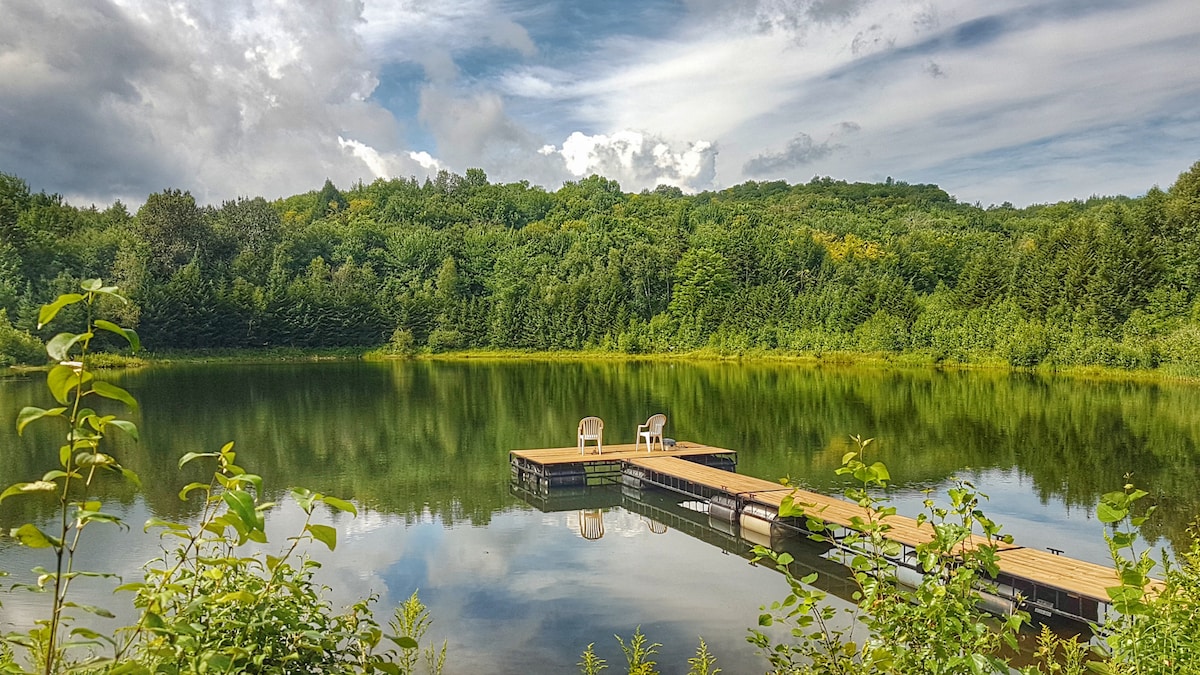 A serene private lake is surrounded by greenery, reflecting the sky and trees. A wooden dock extends into the water, featuring two white chairs positioned for relaxation. The tranquil atmosphere invites outdoor activities and moments of calm.