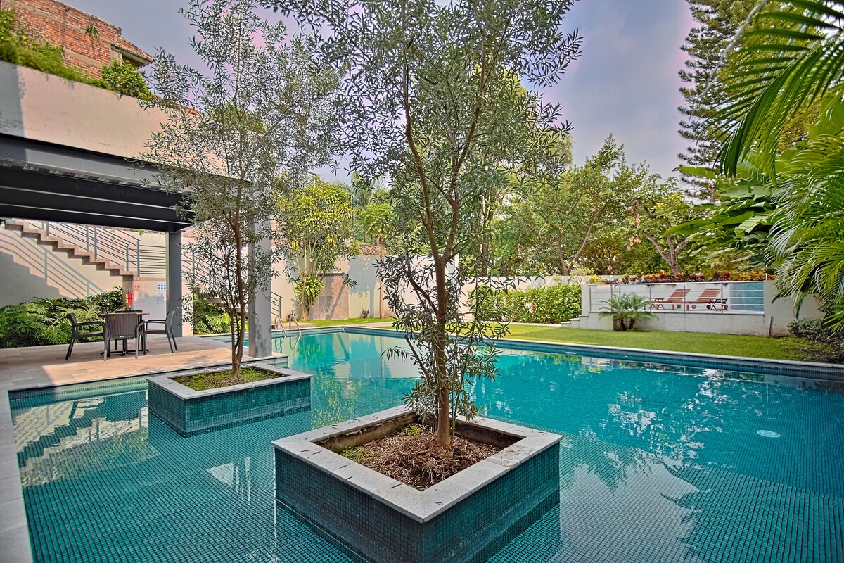 The image displays a serene swimming pool surrounded by lush greenery. Two small trees are situated in planters within the pool, and a spacious lawn is visible in the background. The area is designed for relaxation, with lounge chairs accessible nearby.