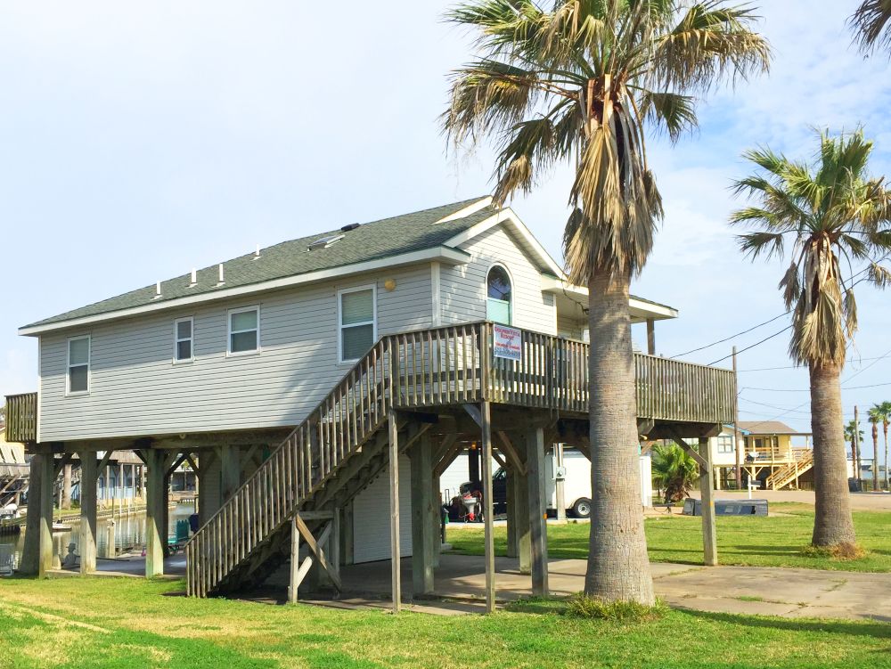 A two-story resort house is elevated on wooden stilts, providing ample parking space underneath. Surrounding palm trees enhance the outdoor area, while large windows reflect natural light. The exterior is finished in light-colored siding, contributing to a relaxed coastal ambiance.