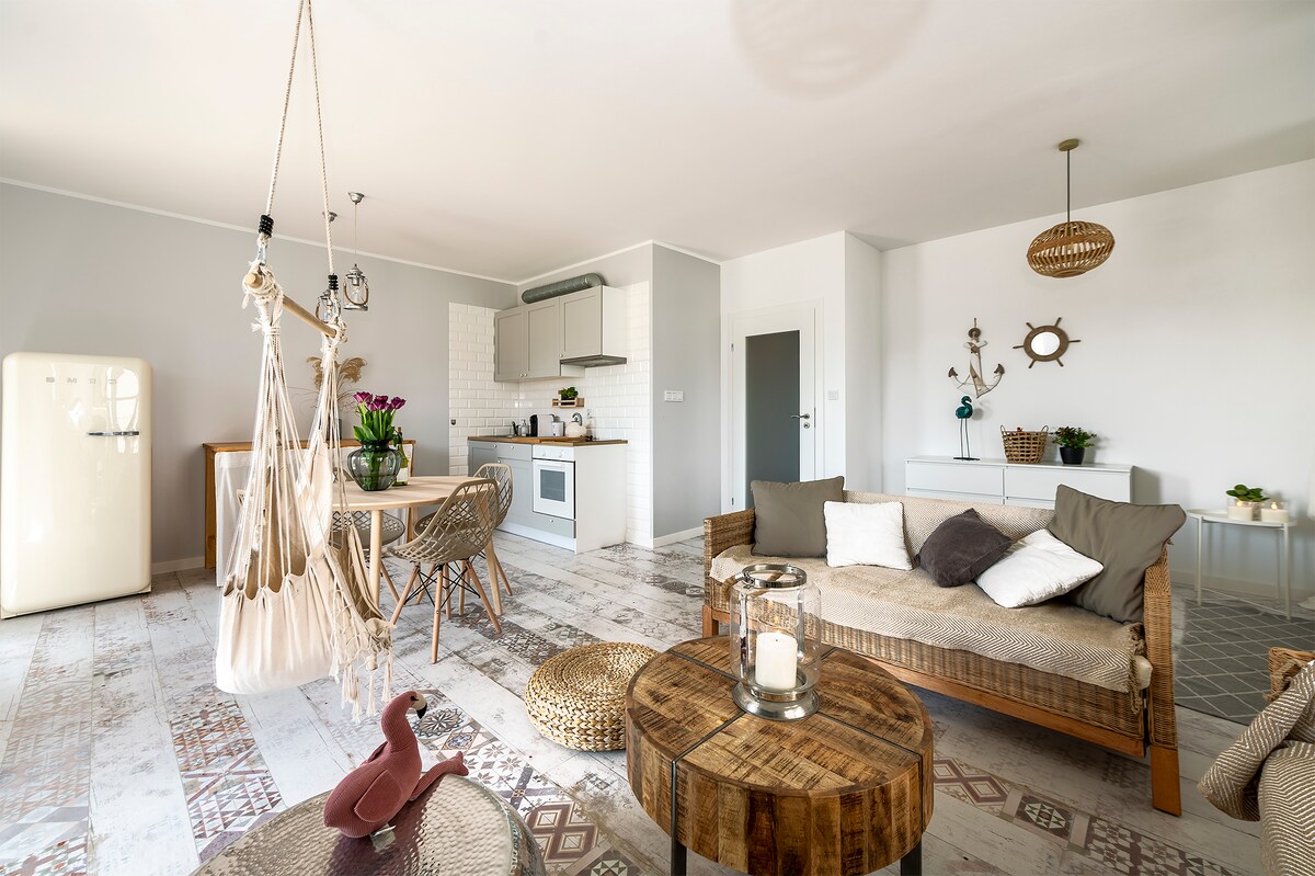 A bright indoor space features a cozy seating area with a woven coffee table and comfortable cushions. A dining table with wooden accents is visible, alongside a stylish kitchen fitted with modern appliances. A hanging chair adds a playful touch to the boho-inspired decor.