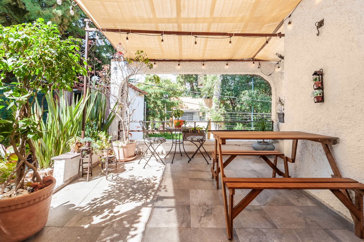 A covered terrace area features a wooden table and chairs, surrounded by potted plants and greenery. Natural light filters through the canopy, creating a serene spot for relaxation. The space is framed by the lush surroundings, offering a peaceful outdoor atmosphere.