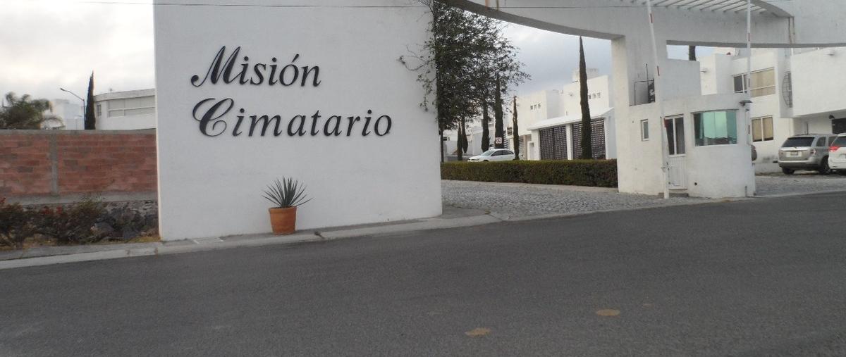 The entrance to a gated community is marked by a large sign that reads 'Misión Cimatario.' A well-maintained pathway leads into the community, flanked by neatly trimmed hedges and decorative plants. The area is peaceful and organized, contributing to a welcoming ambiance.