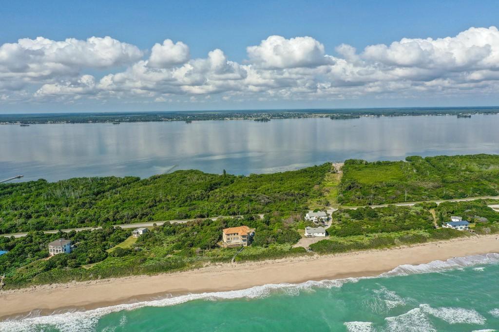 An aerial view showcases the property along a serene beach, with soft waves lapping against the shore. Lush greenery lines the coastline, complemented by a coastal road. The tranquil waters reflect the clouds overhead, creating a peaceful atmosphere in this natural setting.
