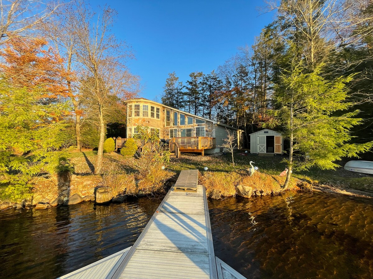 A lakeside view showcases a spacious house with large windows, providing ample natural light. A wooden deck extends from the rear, leading to a dock. Surrounding trees create a private setting, while the calm water reflects the clear sky.