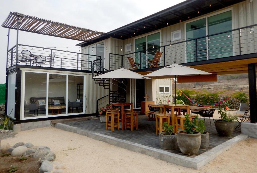 A modern container home is showcased with a spacious outdoor dining area featuring wooden tables and chairs. A covered terrace is seen above, adorned with lounge chairs. The scene is completed with potted plants and hammocks, creating a relaxing outdoor environment.