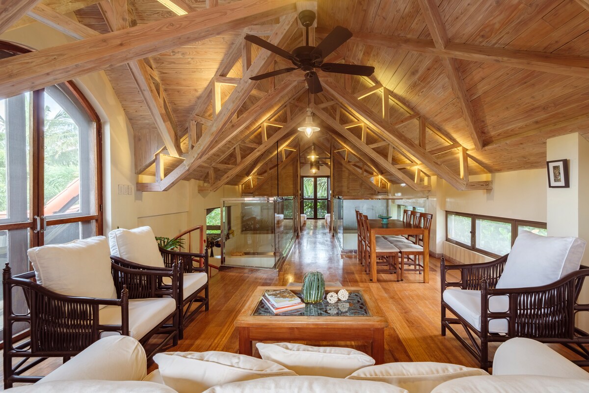 A spacious loft area features tall wooden beams and a ceiling fan, complemented by natural light from large windows. Comfortable seating arrangements with white cushions surround a coffee table, and a dining area is visible in the background, offering a welcoming space for relaxation and gatherings.