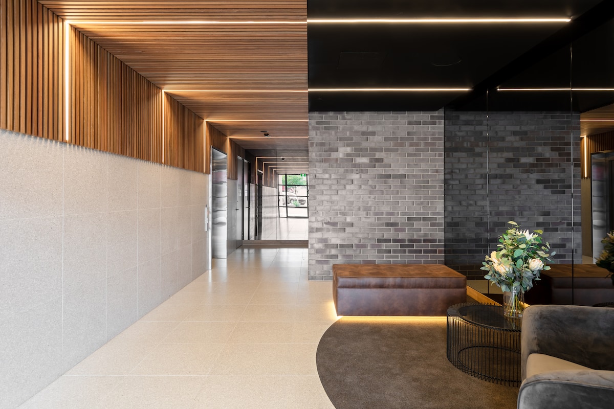The lobby area features a combination of textured wooden panels and smooth stone surfaces. Elegant lighting highlights a communal seating area with plush furniture, while a decorative flower arrangement adds a touch of freshness. Glass doors lead to the exterior, providing a sense of openness.