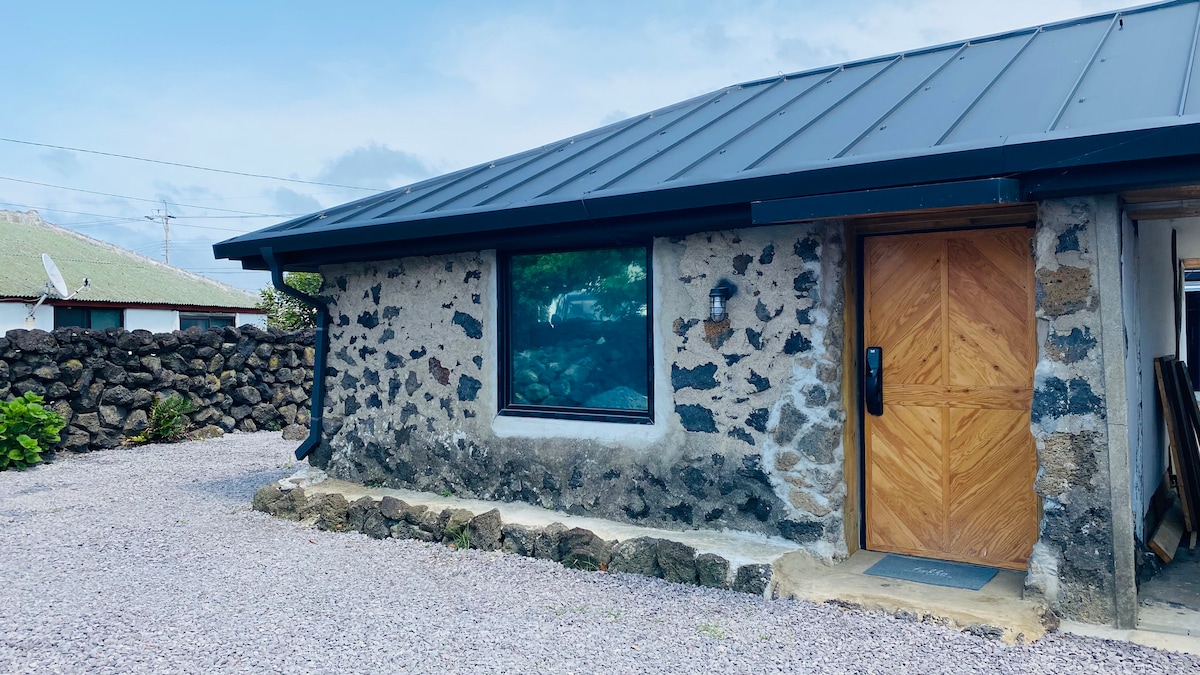 The exterior of a stone building is showcased, featuring a modern metal roof and wooden door. A large window offers a view of the surrounding landscape, while a gravel pathway leads to the entrance. Natural stone walls create a rustic appearance.