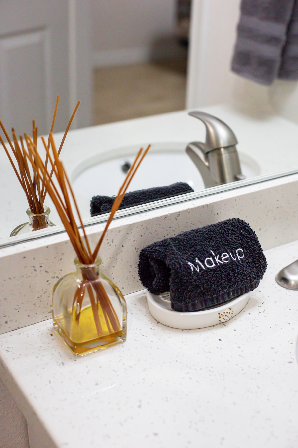 A modern bathroom countertop is displayed, featuring a sleek faucet and a white sink. A glass container holds decorative reed diffusers, while a dark towel with the word 'wake up' is neatly arranged on a small dish. The partially visible mirror adds depth to the scene.