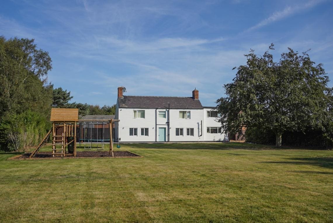 A large, renovated farmhouse is positioned in a spacious green yard, surrounded by trees. On the left, a children's play area with a swingset and climbing frame complements the outdoor space, while the inviting exterior of the farmhouse is highlighted by a clear blue sky.