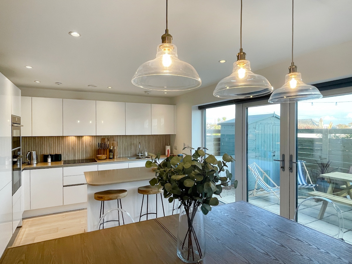 A modern kitchen is equipped with sleek white cabinetry and stainless steel appliances. Wooden bar stools surround a minimalistic island. Three glass pendant lights illuminate the space, complementing the natural light from the large sliding doors that lead to the outdoor area.