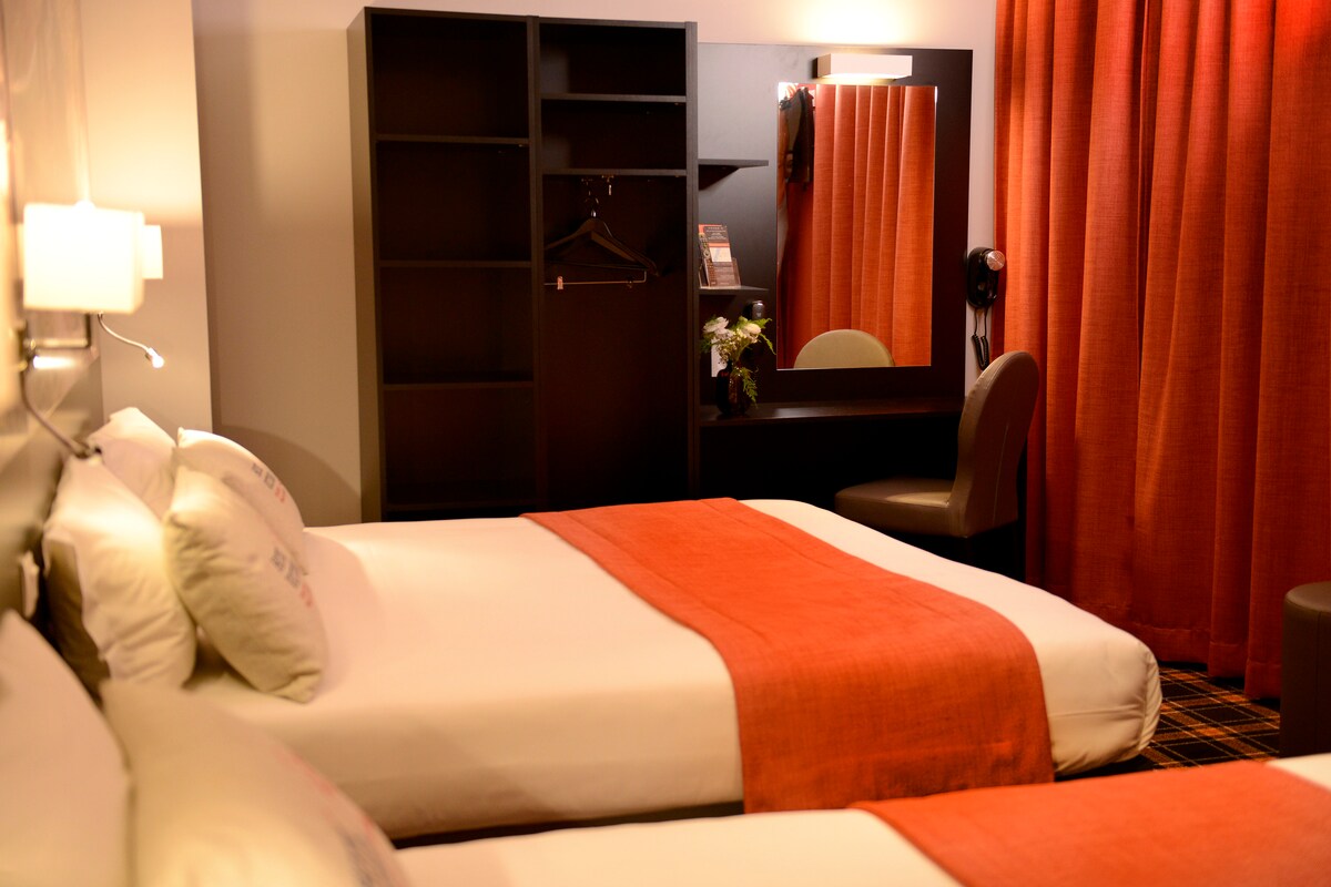 A well-appointed room features a spacious bed and a single bed, accented with an orange blanket. A dark wooden wardrobe and a mirror with vanity lights are visible, alongside a comfortable chair. Bright orange curtains frame the window, allowing natural light into the space.