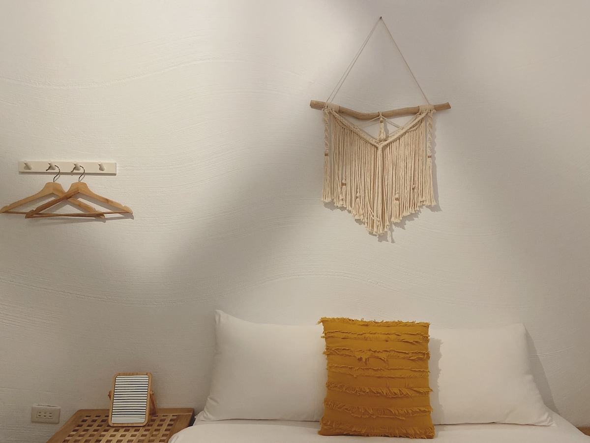A minimalistic bedroom features a spacious bed with white linens, complemented by a mustard-colored decorative pillow. A textile wall hanging adds texture above the bed. A wooden shelf is mounted on the wall with two hangers, offering storage options.